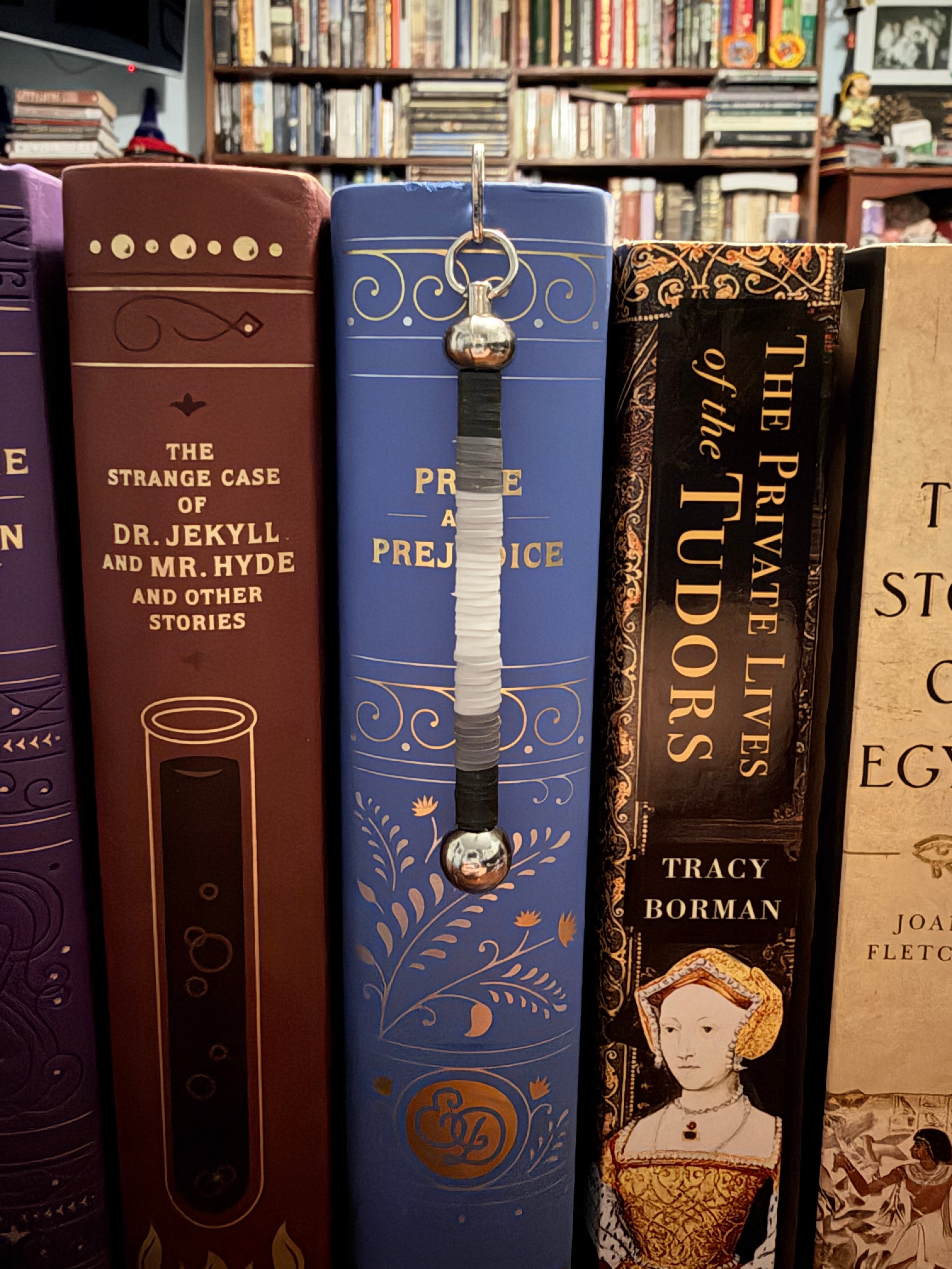 A close-up of a decorative book spine bookmark hanging from a ring on a blue book in a library or bookstore setting, surrounded by other books.