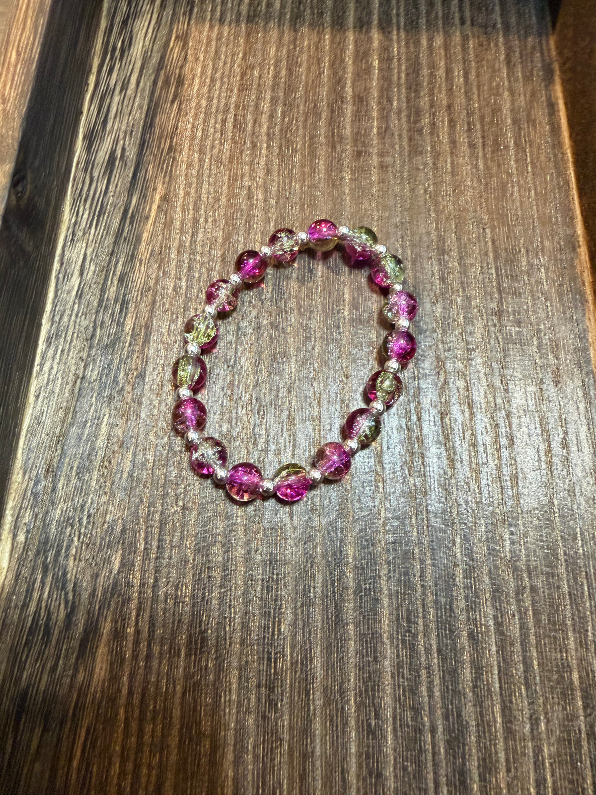 A beaded stretch bracelet with pink, silver, and light green crackle glass beads, displayed on a dark wood surface.