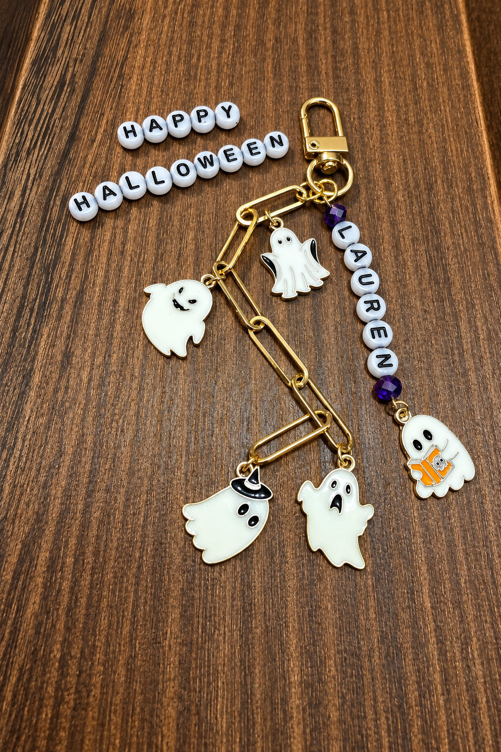 Halloween-themed keychain with gold-tone hardware, white ghost charms, purple beads, and a beaded 'Lauren', displayed on a wooden surface next to a 'Happy Halloween' bead garland.