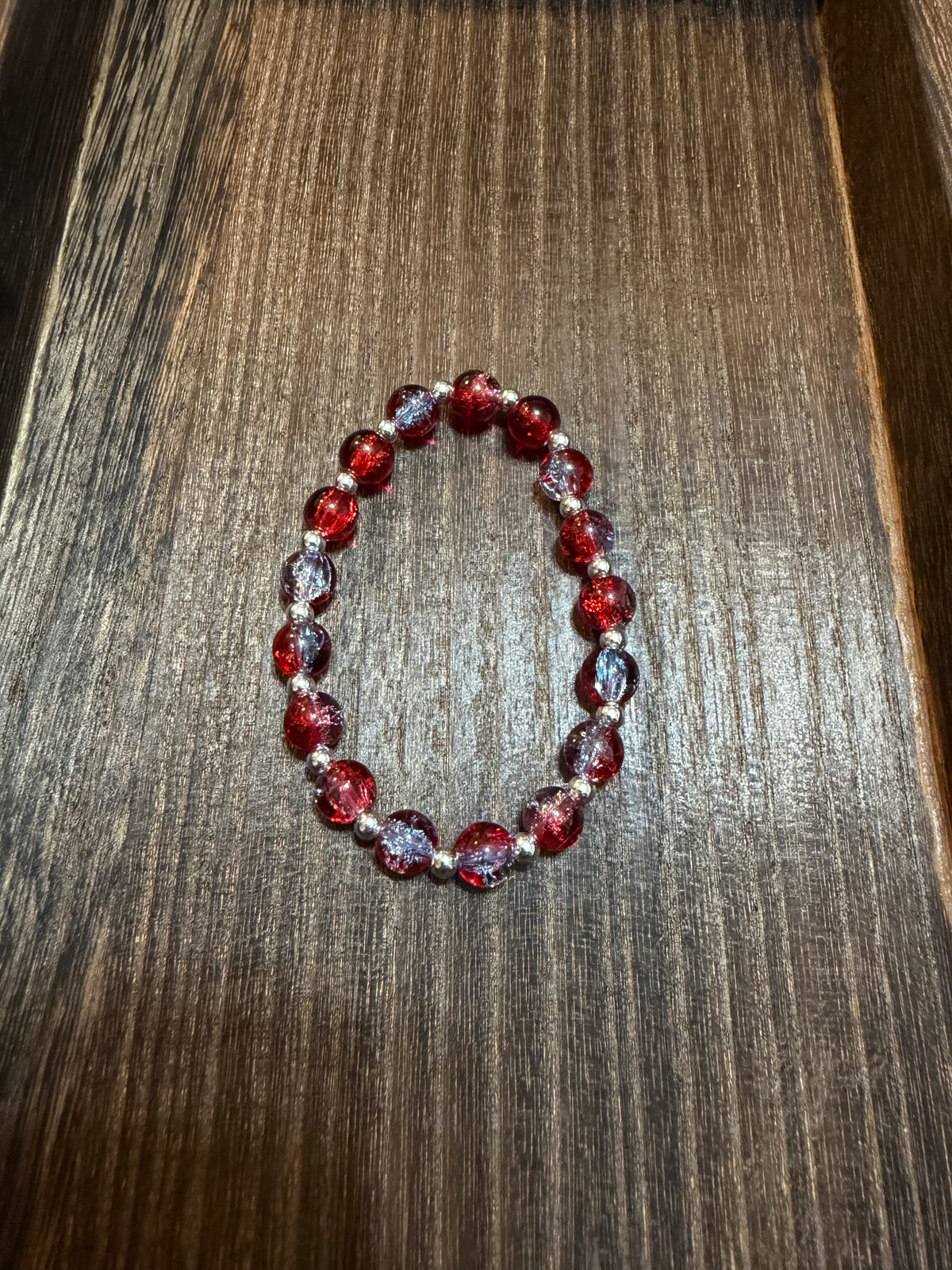 A beaded stretch bracelet made of crackle glass beads in red and light blue with silver spacer beads, displayed on a dark brown wooden surface.