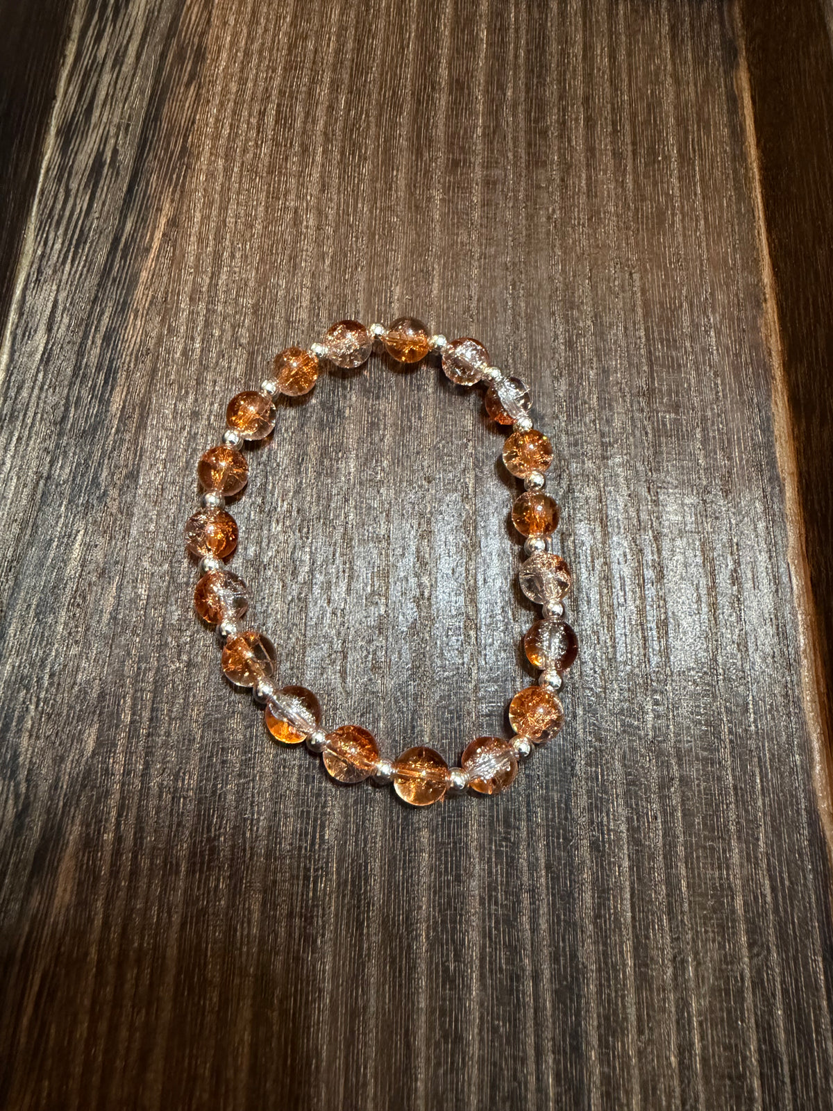 A beaded stretch bracelet made of amber-orange and silver crackle glass beads, displayed on a dark brown wood-grain surface.