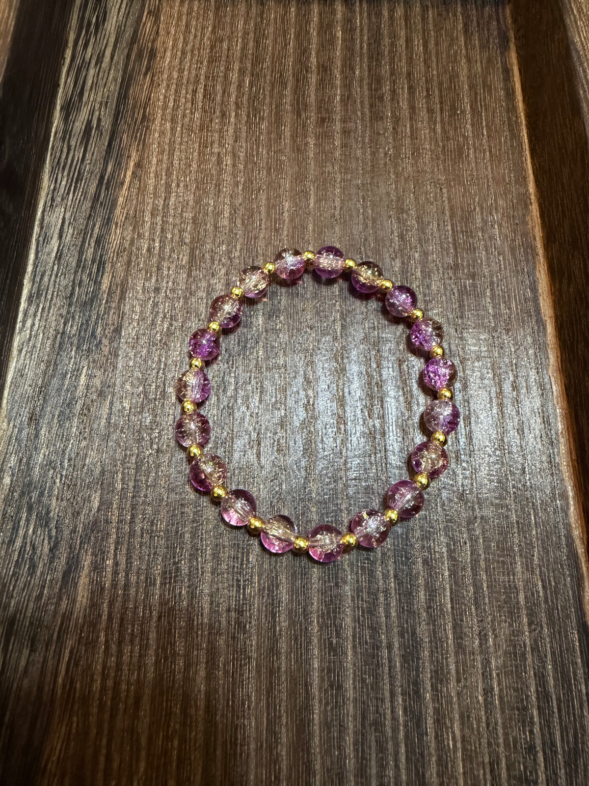 A beaded stretch bracelet with lavender, pink, and gold crackle glass beads separated by small gold spacers, displayed on a dark brown wooden surface.