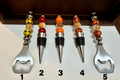 Five bottle openers and wine corks arranged upright on a white surface, with two featuring metal bottle opener ends and three featuring wine corks. The stoppers have colorful decorative beads in yellow, orange, red, black, and white, with some showing a small pumpkin pattern.