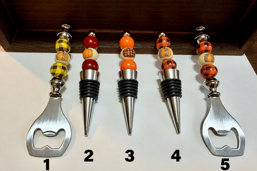Five bottle openers and wine corks arranged upright on a white surface, with two featuring metal bottle opener ends and three featuring wine corks. The stoppers have colorful decorative beads in yellow, orange, red, black, and white, with some showing a small pumpkin pattern.