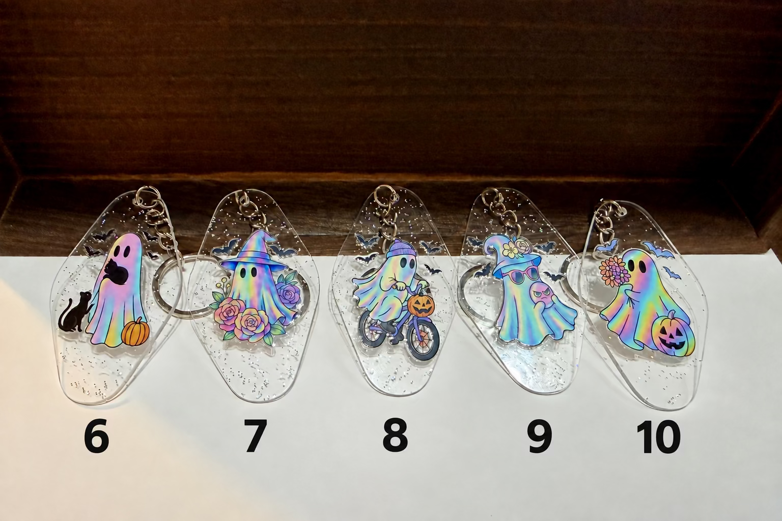 Five clear acrylic keychains with colorful ghost-themed charms are lined up on a white surface against a dark wood background.