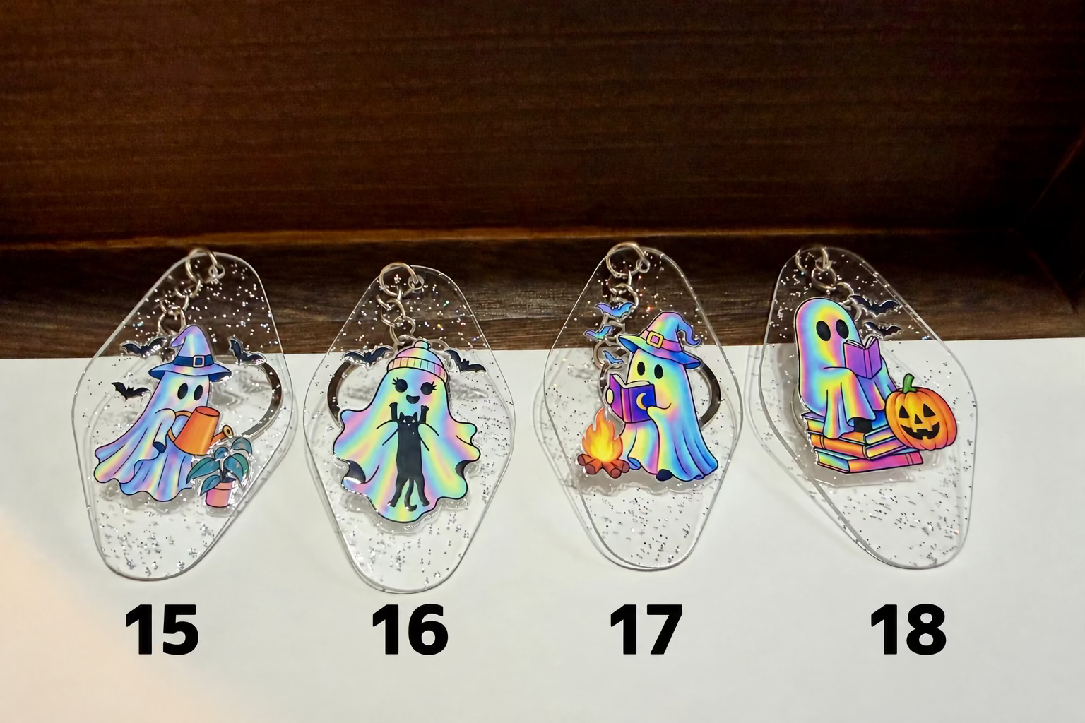 Four clear, glittery acrylic keychains shaped like ghosts with Halloween-themed designs, displayed on a white surface against a dark wood background.