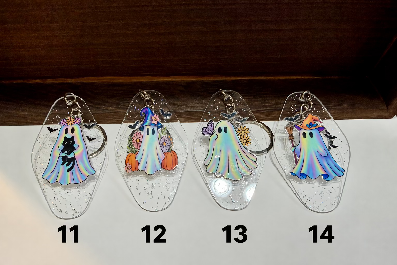 Four clear acrylic keychains with glittery ghost designs in different Halloween-themed colors, displayed on a white surface with a dark wood background.