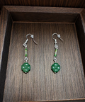 A pair of handmade dangle earrings with green carved beads and silver hooks, displayed on a dark wooden tray.