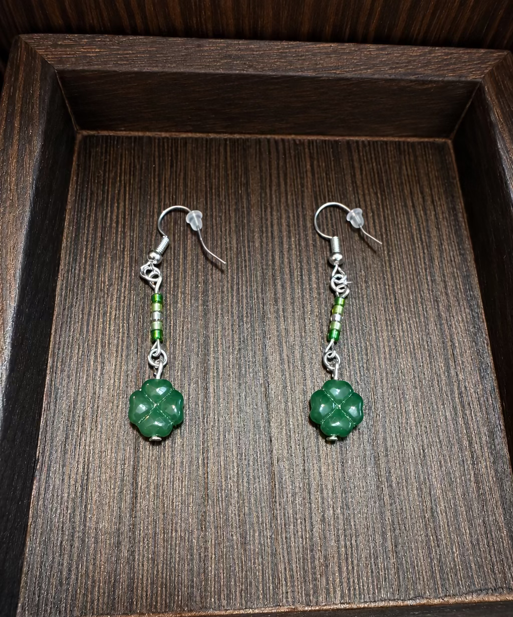 A pair of handmade dangle earrings with green carved beads and silver hooks, displayed on a dark wooden tray.
