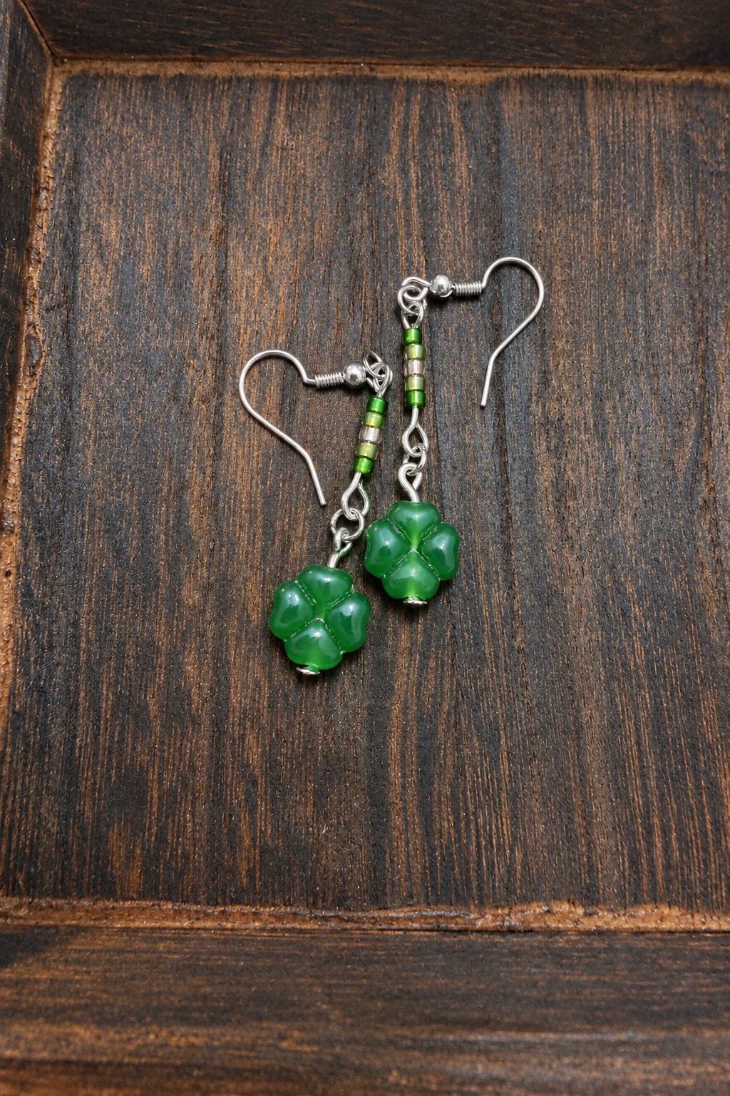A pair of handmade dangle earrings featuring green clover-shaped beads suspended from silver wire hooks with small green, light green, and gold beads, displayed on a dark wooden surface.