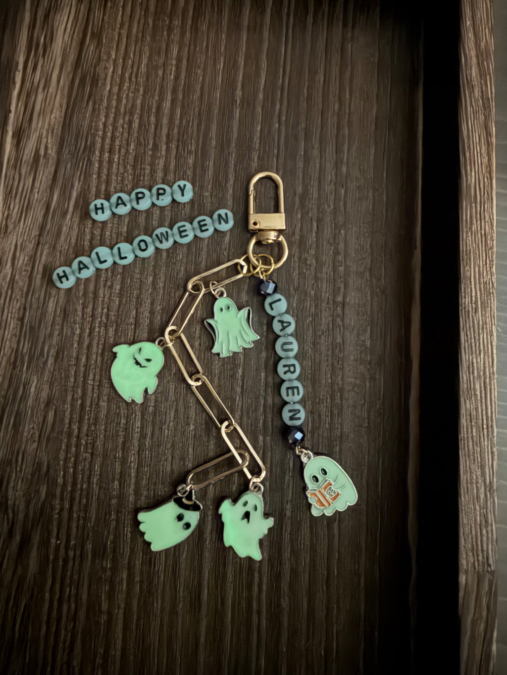 A Halloween-themed keychain or bag charm with a gold-tone clasp, paperclip-style links, a gold-toned bead chain, and multiple ghost charms in pale mint green, dark blue beads, and a small orange 'H' accent, displayed on a dark wood surface with 'HAPPY HALLOWEEN' letter tiles nearby.