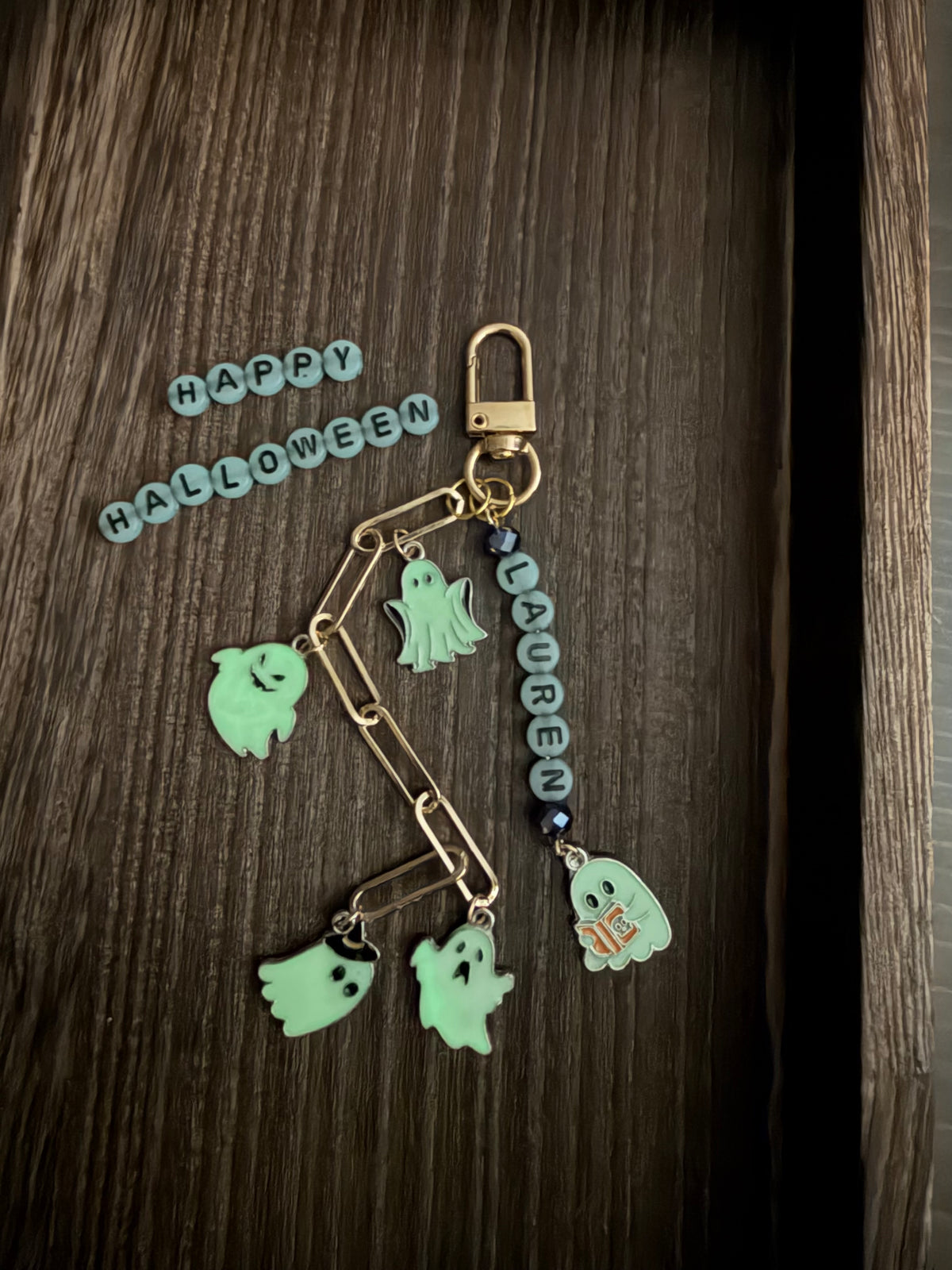 A Halloween-themed keychain or bag charm with a gold-tone clasp, paperclip-style links, a gold-toned bead chain, and multiple ghost charms in pale mint green, dark blue beads, and a small orange 'H' accent, displayed on a dark wood surface with 'HAPPY HALLOWEEN' letter tiles nearby.