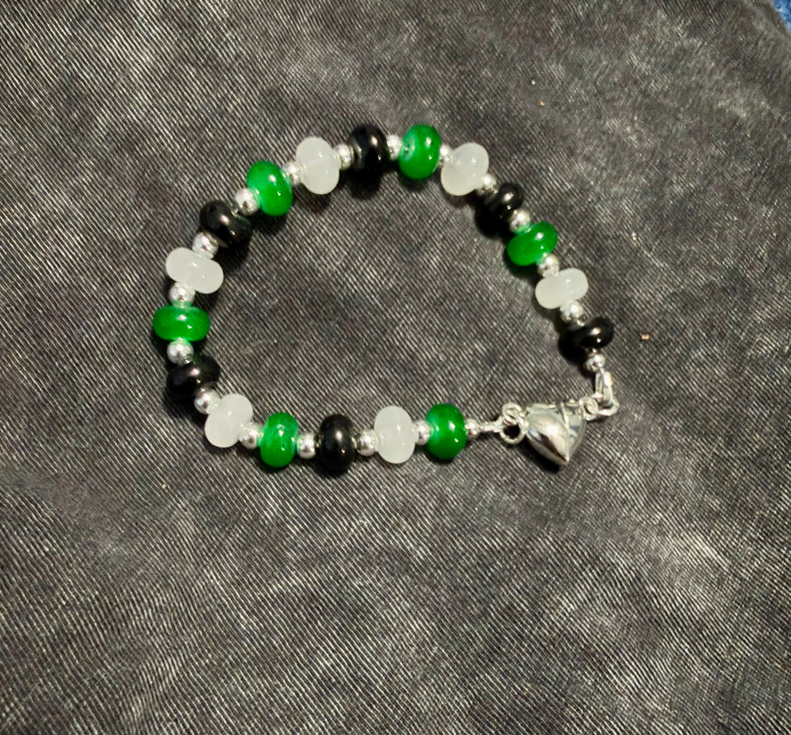 A beaded bracelet with alternating green, white, and black beads, featuring a silver heart clasp, displayed on a dark gray textured fabric background.