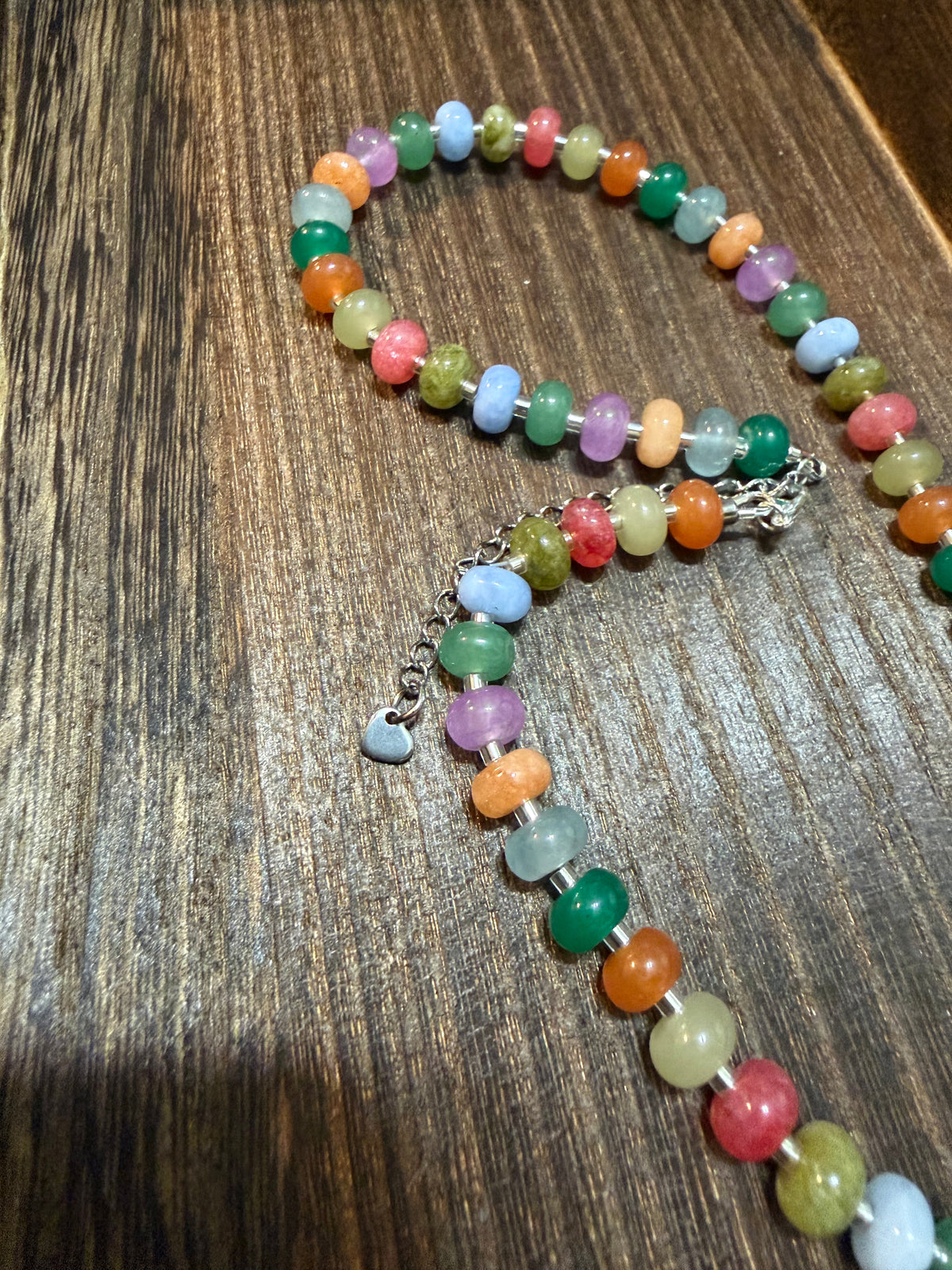 A multicolor beaded necklace with oval polished stones in shades of green, orange, pink, blue, yellow, and purple, displayed on a dark wood surface.