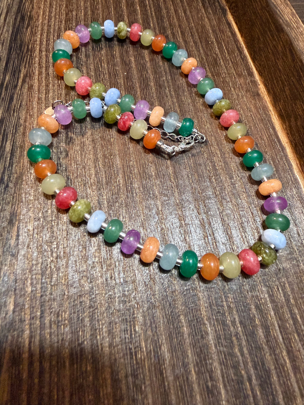 A beaded necklace made of small, colorful polished gemstone beads in multiple shades, arranged on a dark wood surface.