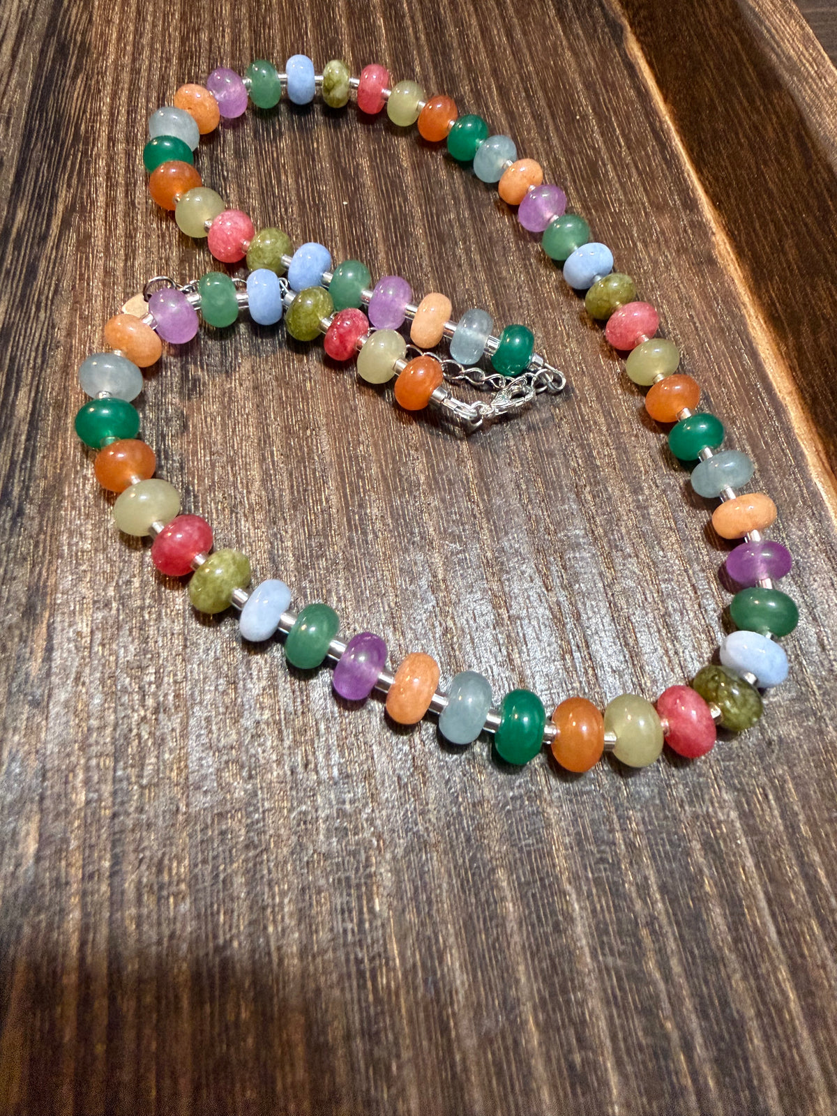A beaded necklace made of small, colorful polished gemstone beads in multiple shades, arranged on a dark wood surface.
