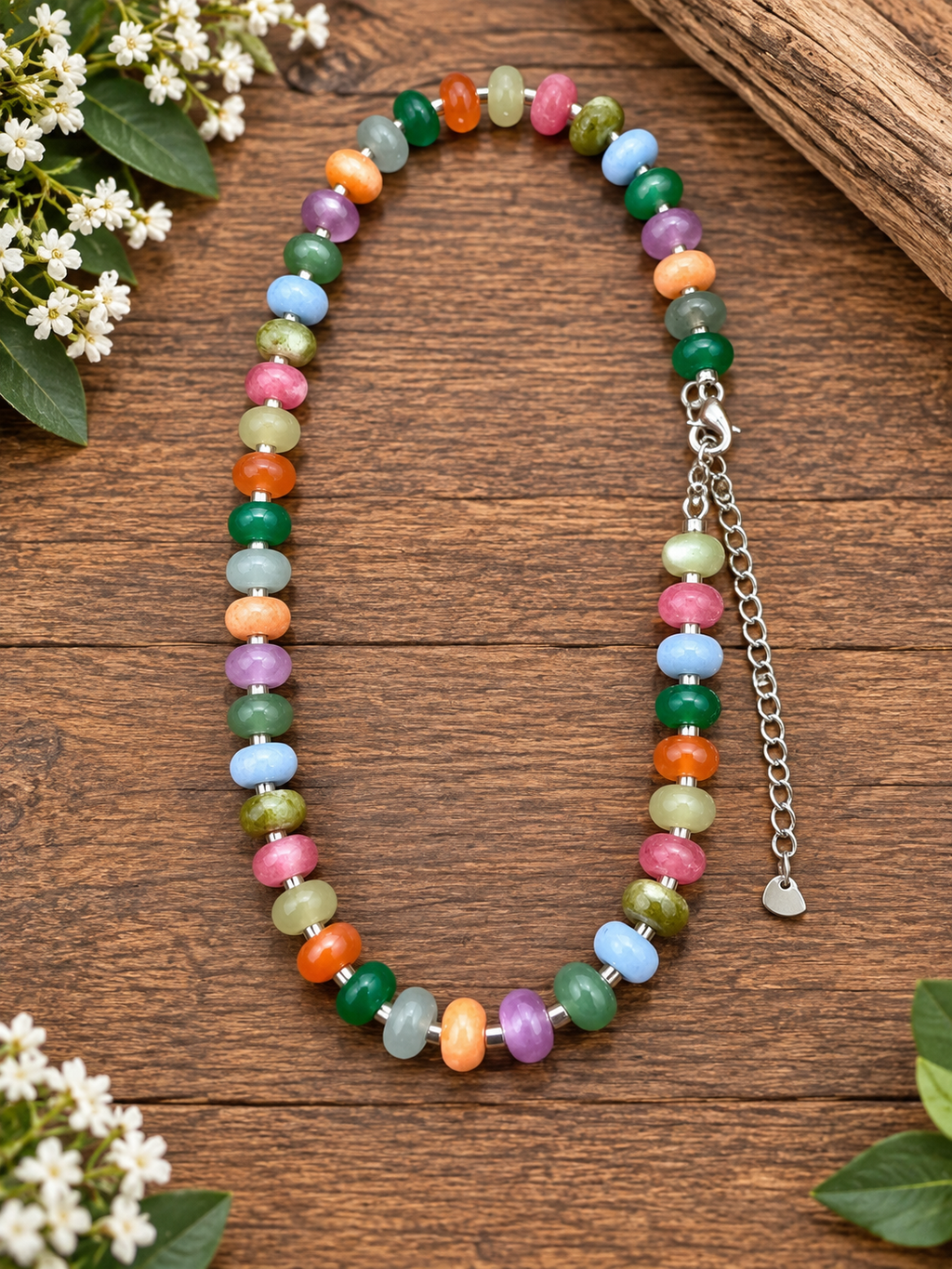A beaded necklace made of colorful rondelle agate gemstone beads in pink, green, orange, blue, purple, and yellow, arranged on a rustic wooden surface with small white flowers and wood accents.