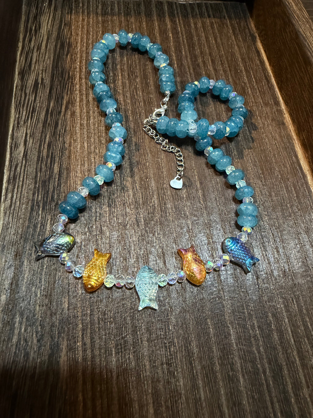 A beaded necklace with light blue stone beads, a silver chain extender, and five colorful fish beads in blue, gold, and iridescent tones, displayed on a dark wood surface.