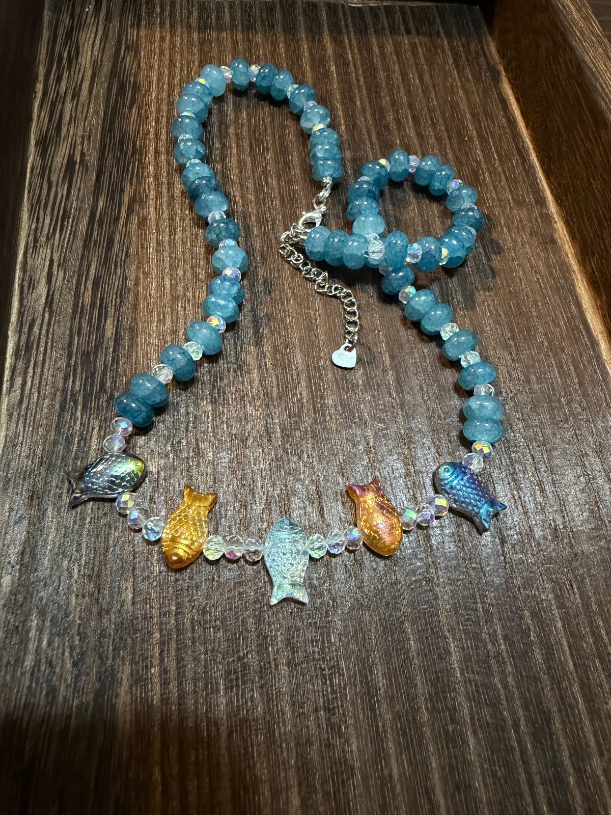 A beaded necklace with light blue stone beads, a silver chain extender, and five colorful fish beads in blue, gold, and iridescent tones, displayed on a dark wood surface.