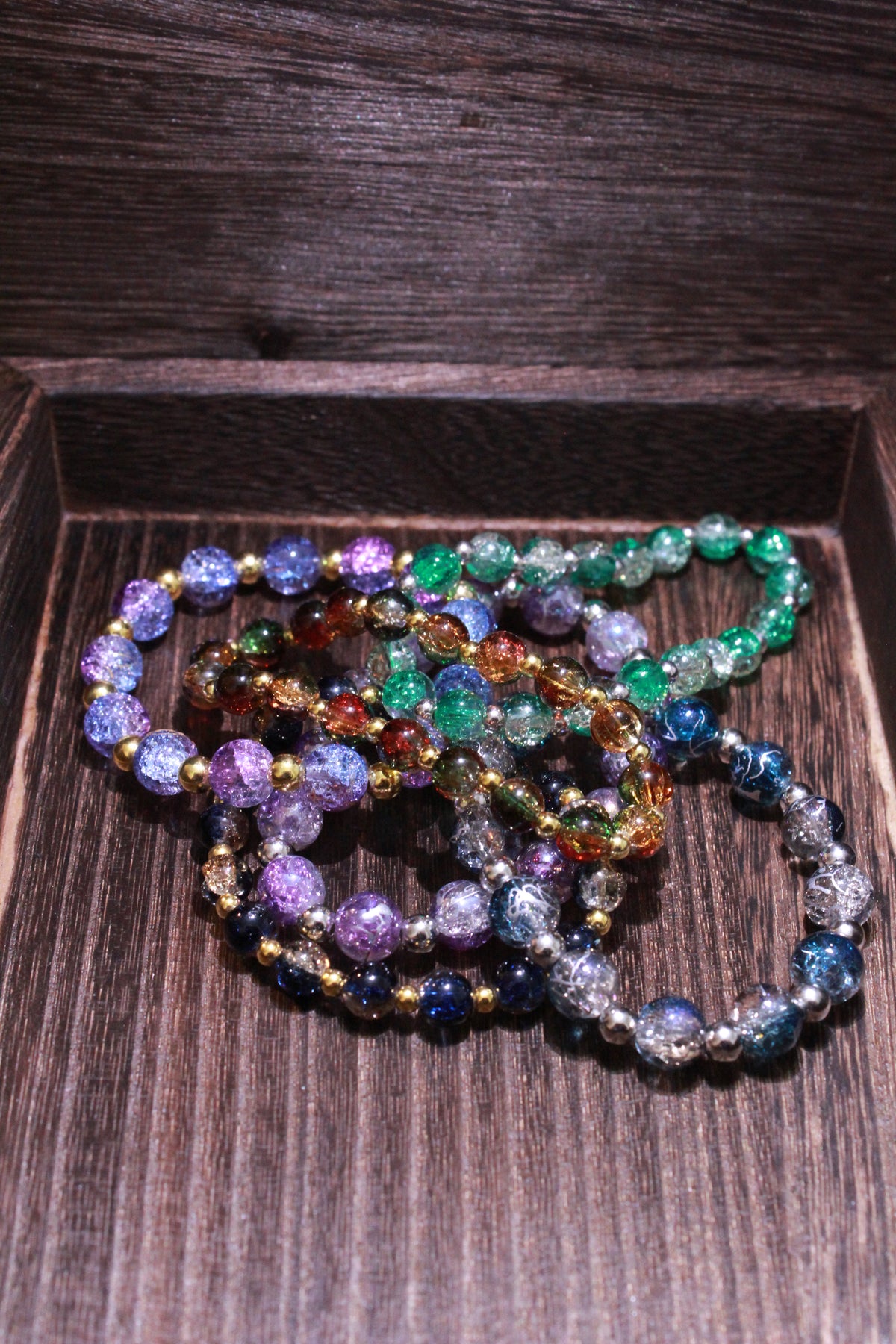 A set of colorful crackle glass beaded bracelets with gold and silver spacers, displayed inside a dark brown wooden box.