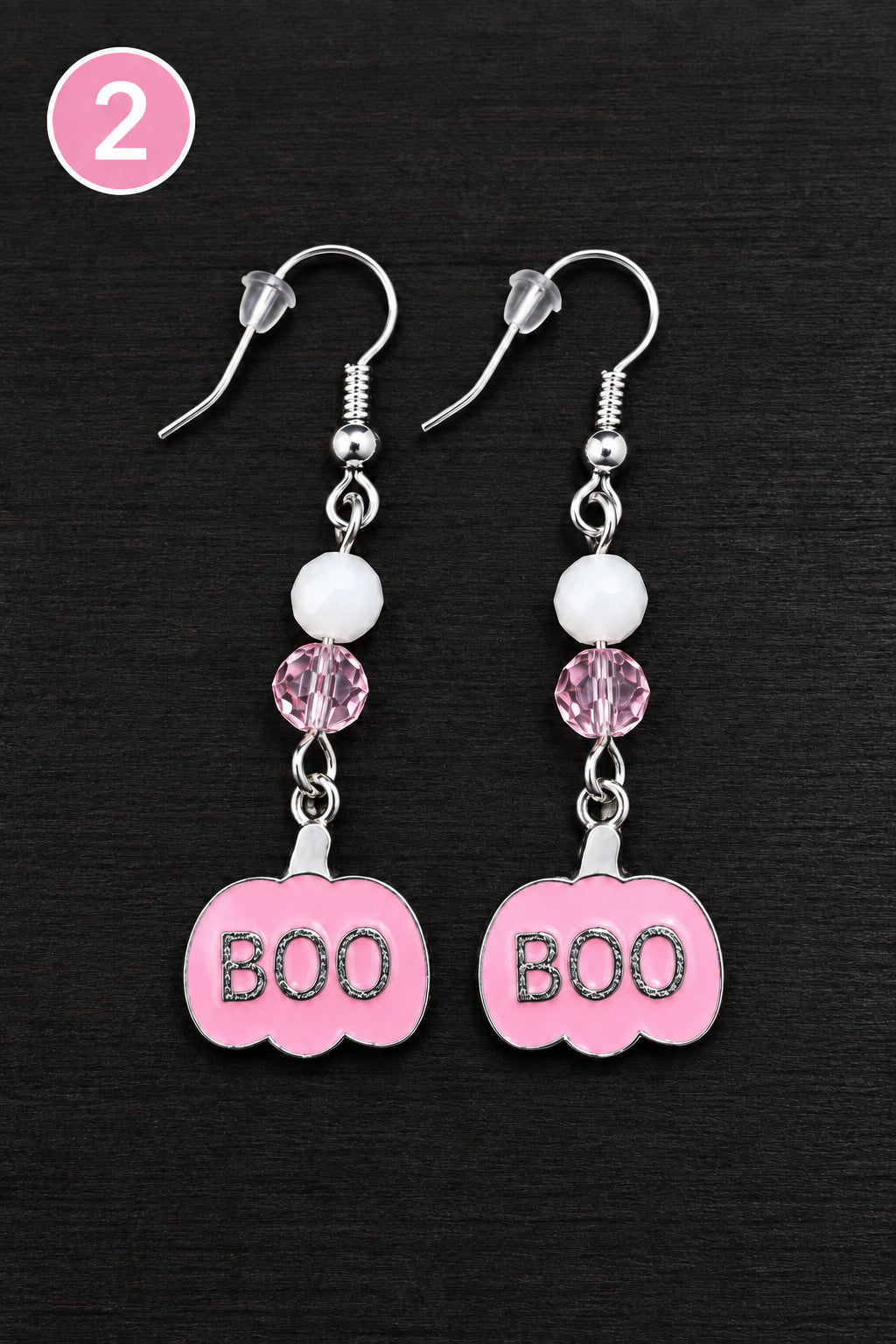 A pair of dangling earrings with silver fishhook wires, white faceted beads, faceted pink beads, and a pink pumpkin-shaped charm with the word 'BOO', displayed on a dark wood surface.