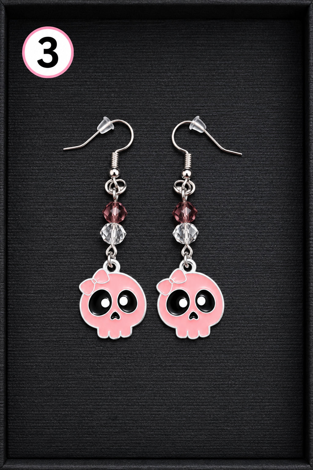 A pair of silver dangle earrings with pink skull charms, displayed on a textured black background. Each earring features a silver fishhook, a small faceted burgundy bead, a clear faceted bead, and a pink skull charm with black eyes.