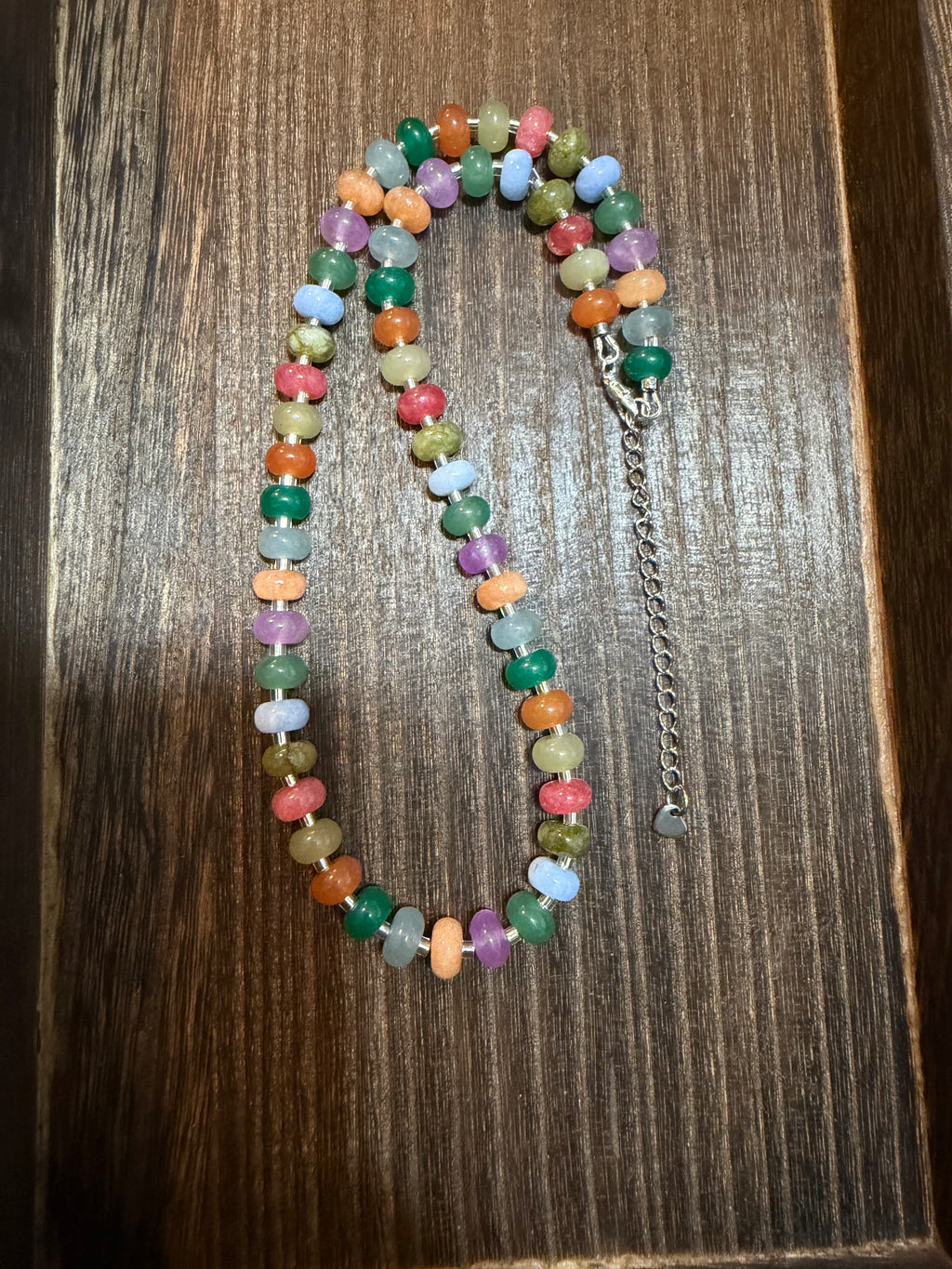A multicolor beaded necklace made of small round gemstone beads in various colors, laid out on a dark wood surface. It features a silver-tone lobster clasp and an adjustable silver-tone chain extender.