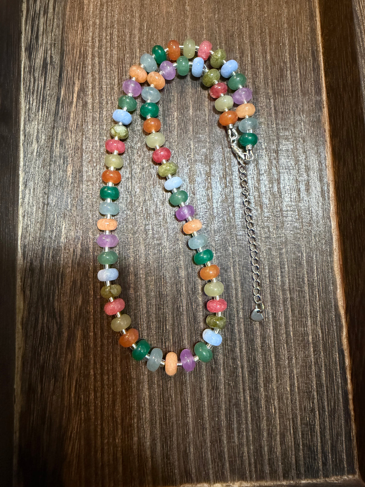 A multicolor beaded necklace made of small round gemstone beads in various colors, laid out on a dark wood surface. It features a silver-tone lobster clasp and an adjustable silver-tone chain extender.