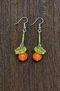 A pair of handmade dangle earrings featuring green glassy leaves and carved orange stone pumpkins, suspended from silver fishhook wires and three shades of green seed beads displayed on a dark wood surface.