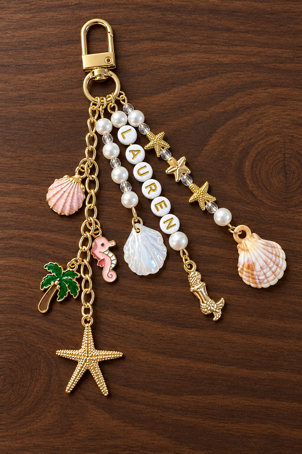 A gold-tone keychain bag charm featuring a lobster clasp and multiple dangling charms including seashells, a starfish, a palm tree, a pink mermaid figure, and letter beads spelling 'LAUREN' on a wooden surface.
