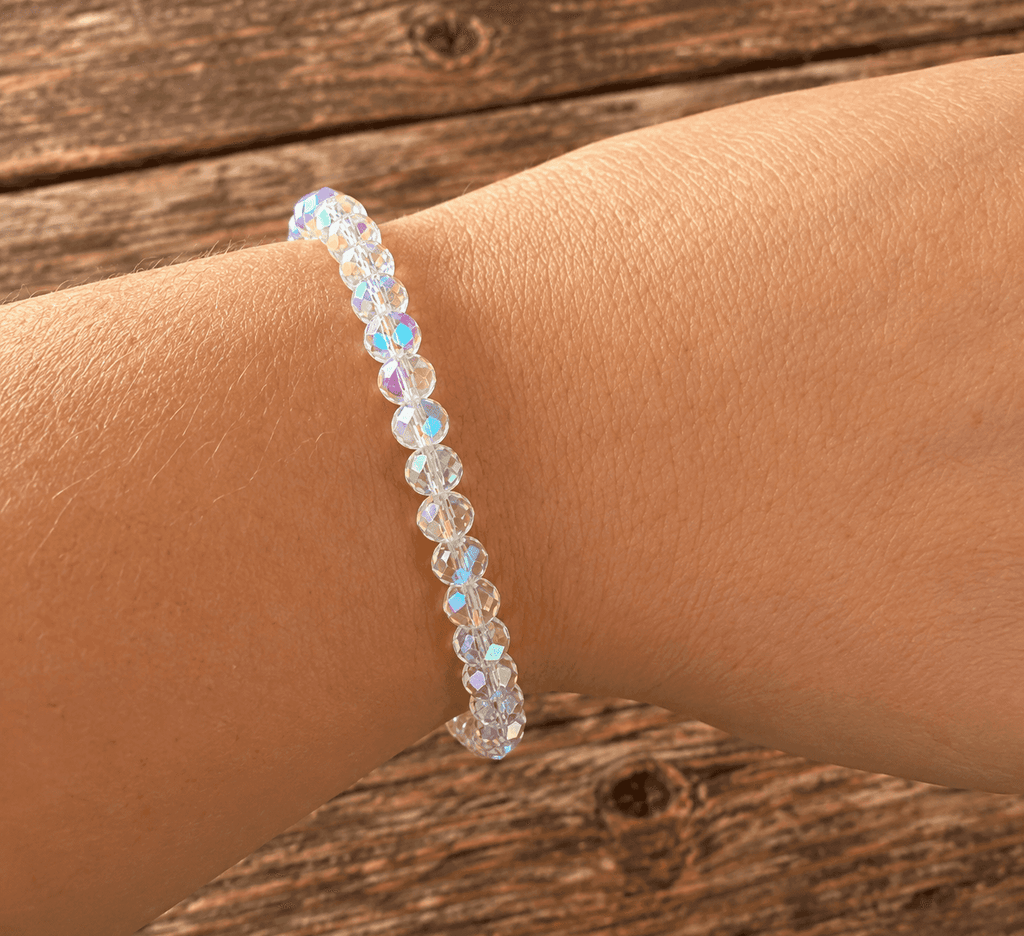 A beaded stretch bracelet with iridescent, rainbow-flecked faceted beads worn on a person's wrist, set against a rustic wooden plank background.