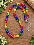 A beaded necklace made of colorful round beads in rainbow colors, arranged on a wooden surface with small white flowers and green leaves around it. The necklace has a silver-tone clasp and chain extender with a small heart charm.