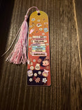 A decorative holographic acrylic bookmark with a matching pink tassel lies on a dark wood surface. The bookmark features illustrated book spines and fantasy-themed labels including 'True Crime', 'Romance', 'Fantasy', and 'Mystery', along with small floral and teacup motifs.