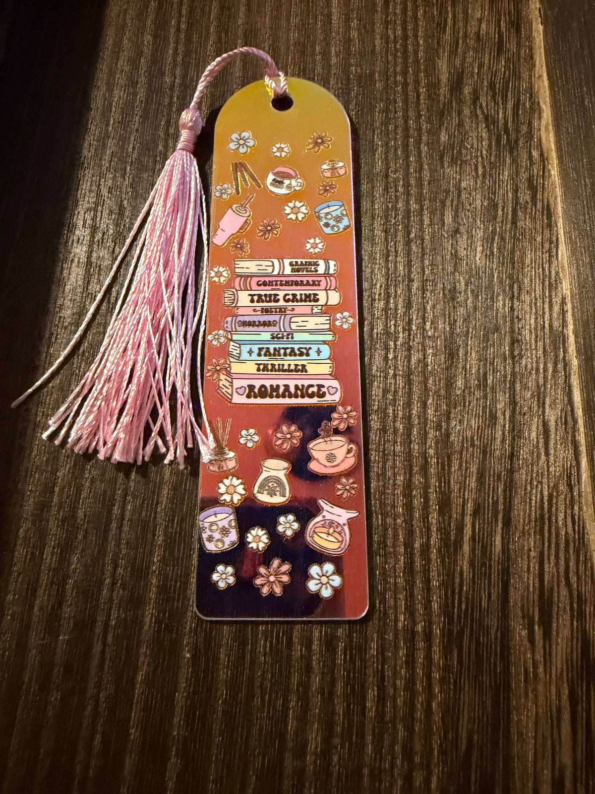 A decorative holographic acrylic bookmark with a matching pink tassel lies on a dark wood surface. The bookmark features illustrated book spines and fantasy-themed labels including 'True Crime', 'Romance', 'Fantasy', and 'Mystery', along with small floral and teacup motifs.