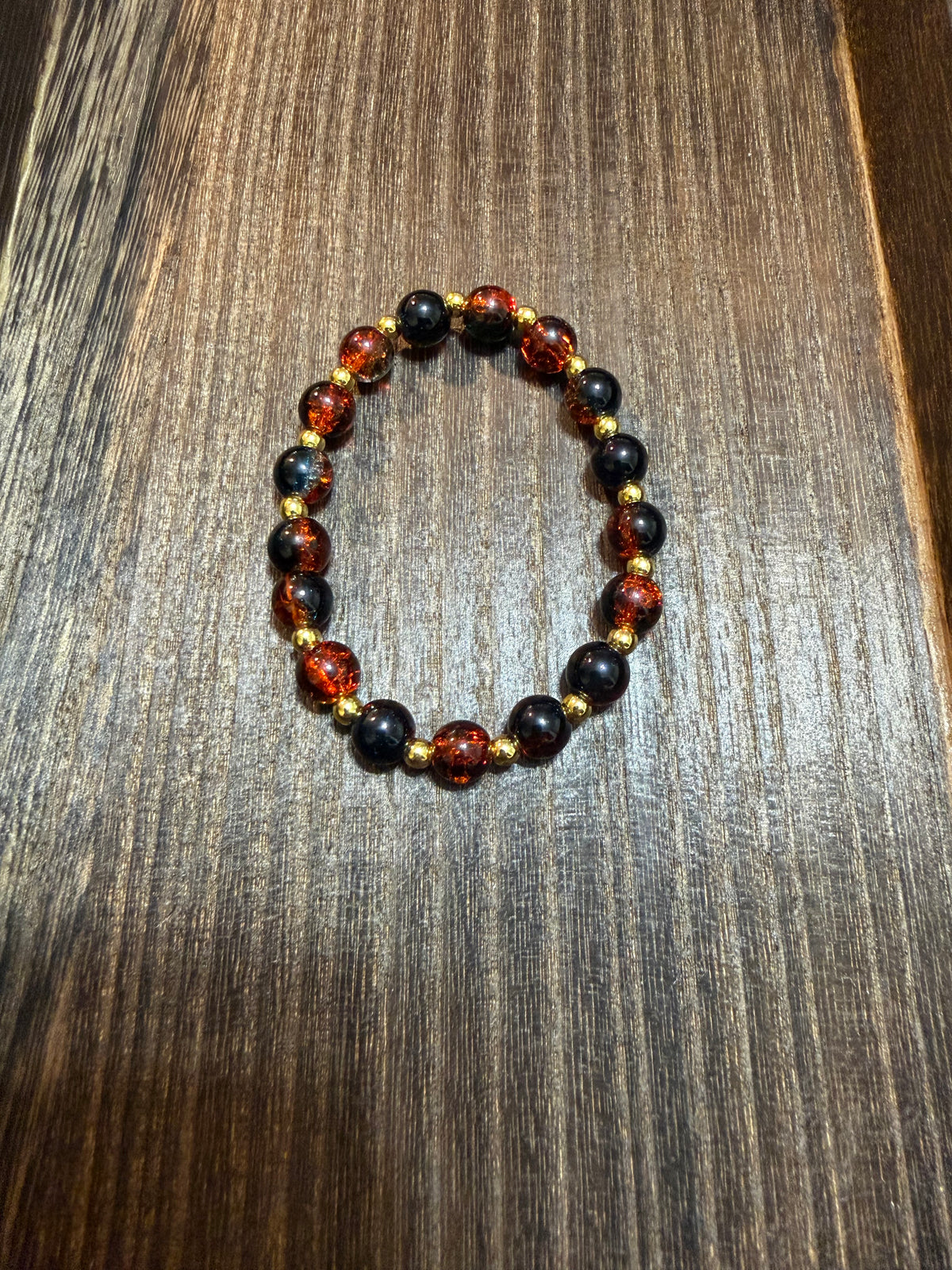 A beaded stretch bracelet featuring deep orange and black crackle glass beads, separated by small gold spacer beads, displayed on a dark wood surface.