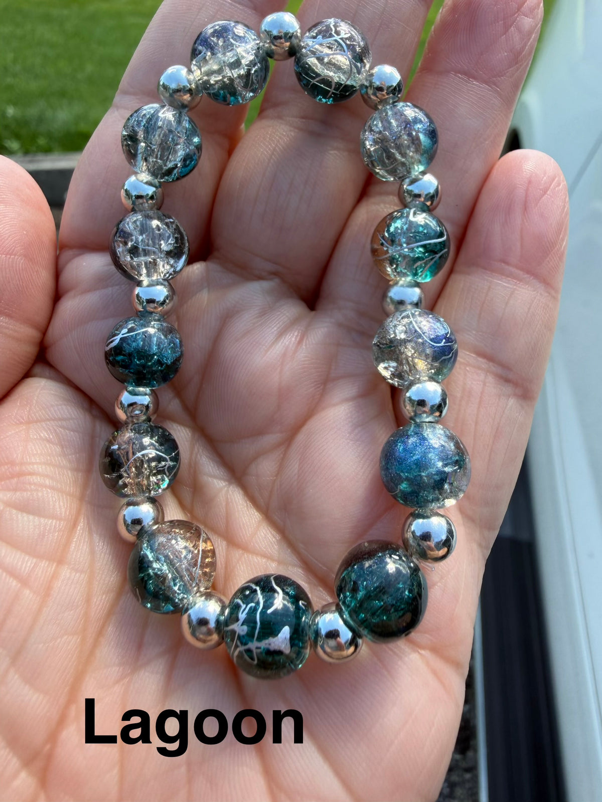 A beaded stretch bracelet labeled 'Lagoon' held in a hand. The bracelet features iridescent blue-green and clear beads with metallic silver accents.