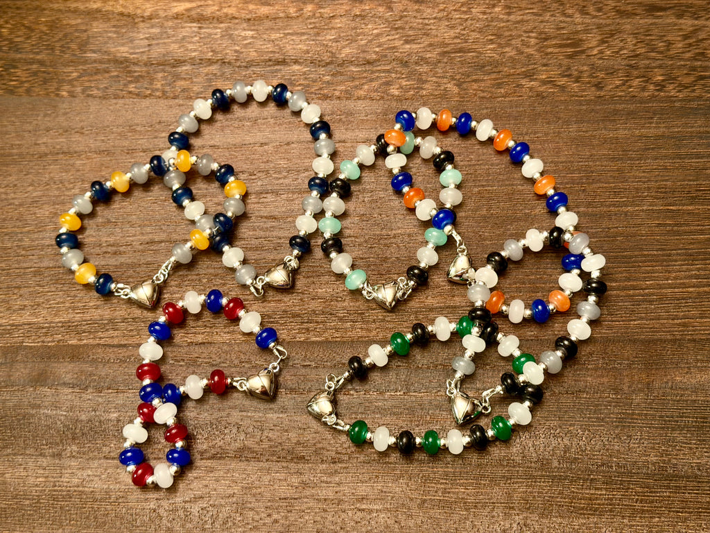 Several beaded bracelets with silver-toned heart clasps arranged on a brown wooden surface. The bracelets feature various bead combinations in blue, white, gold, red, black, green, grey, and orange along with silver spacer beads.