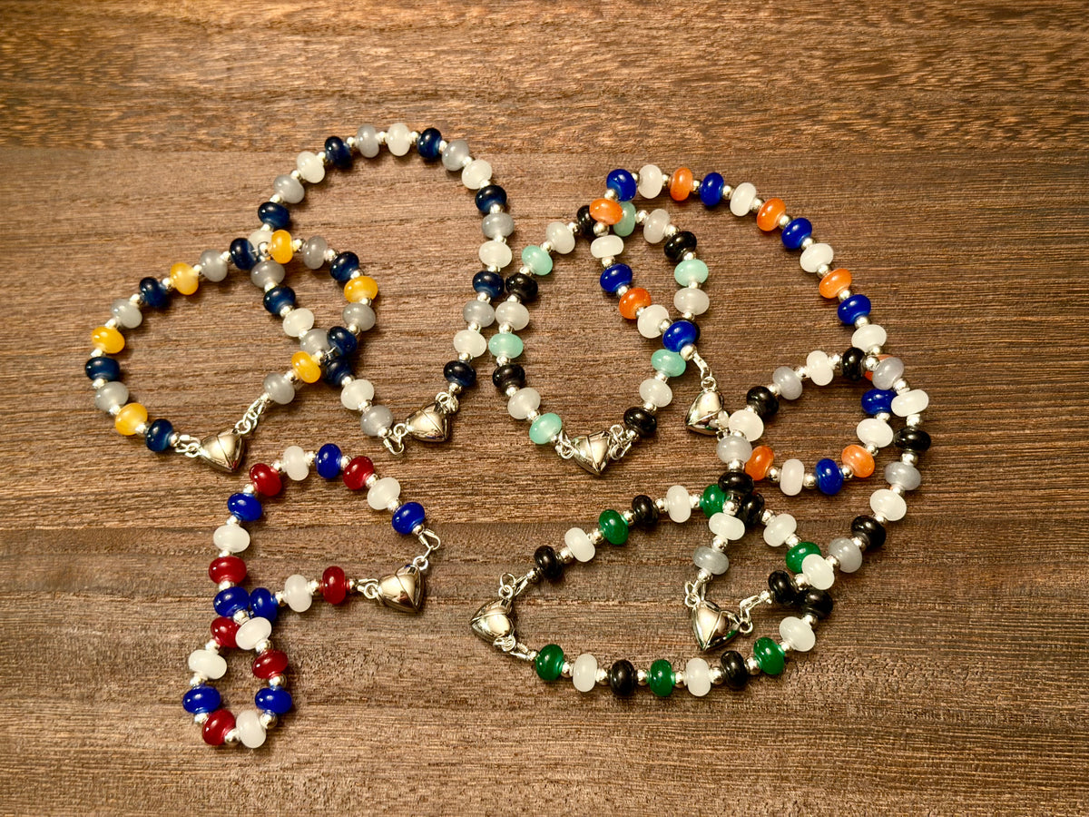 Several beaded bracelets with silver-toned heart clasps arranged on a brown wooden surface. The bracelets feature various bead combinations in blue, white, gold, red, black, green, grey, and orange along with silver spacer beads.
