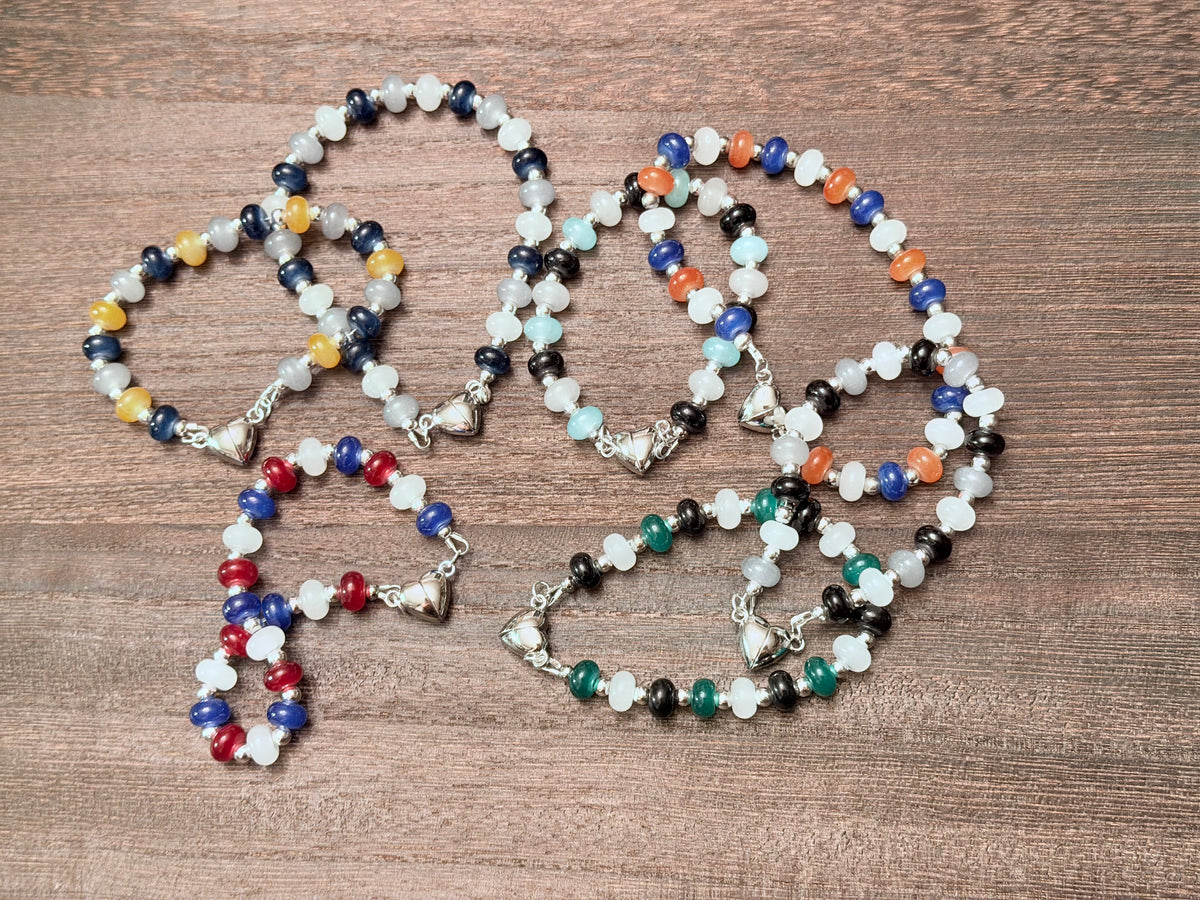 A set of beaded bracelets arranged on a brown wooden surface. The jewelry features multi-colored glass beads in white, blue, red, gold, orange, green, and black, accented with silver-toned metal spacers and a heart shaped magnetic clasp.