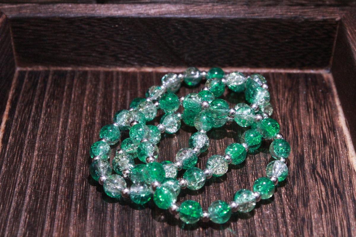 Three beaded bracelets made of crackle glass beads in green and clear with silver separated by small silver spacer beads, displayed on a dark wood surface.