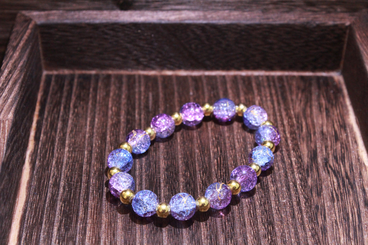 A beaded stretch bracelet featuring crackle purple and blue beads separated by small gold spacer beads, displayed on a dark wood tray.