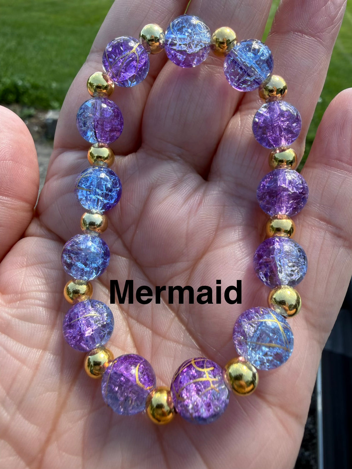 A beaded bracelet with purple and blue crackle glass beads separated by shiny gold spacer beads, held in a person's hand. The word 'Mermaid' is overlaid in black text across the center.