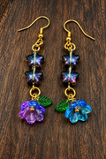 A pair of handmade dangle earrings with gold-tone hooks, dark iridescent beads, green leaves, and purple-blue flower charms, displayed on a dark wood surface.