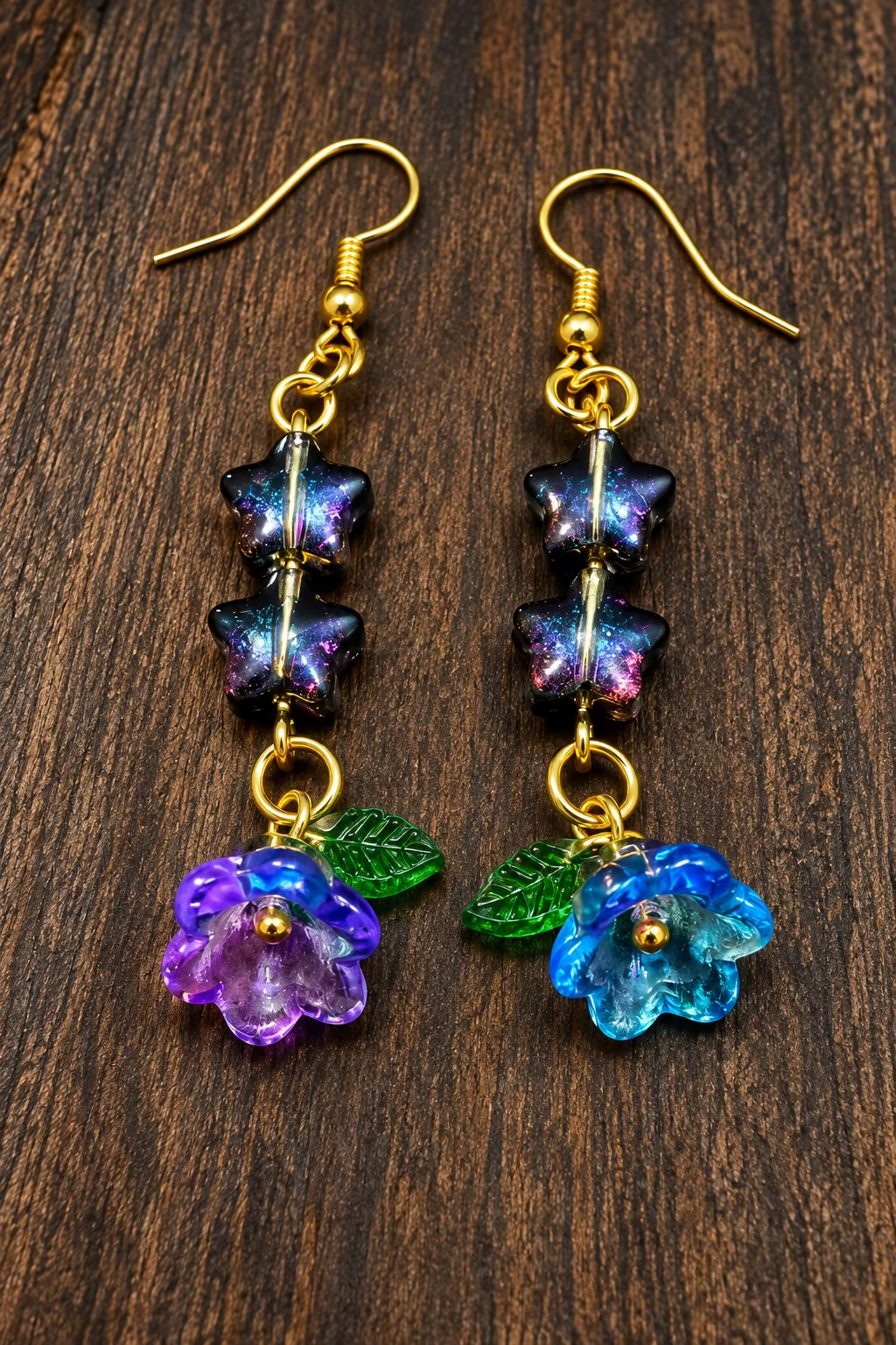 A pair of handmade dangle earrings with gold-tone hooks, dark iridescent beads, green leaves, and purple-blue flower charms, displayed on a dark wood surface.