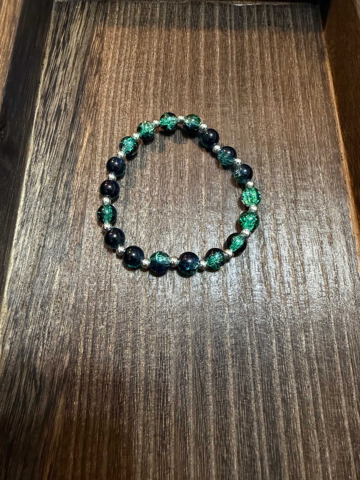 A beaded stretch bracelet featuring green, dark blue, black, and silver crackle glass  beads with small silver spacer beads, displayed on a dark wood surface.