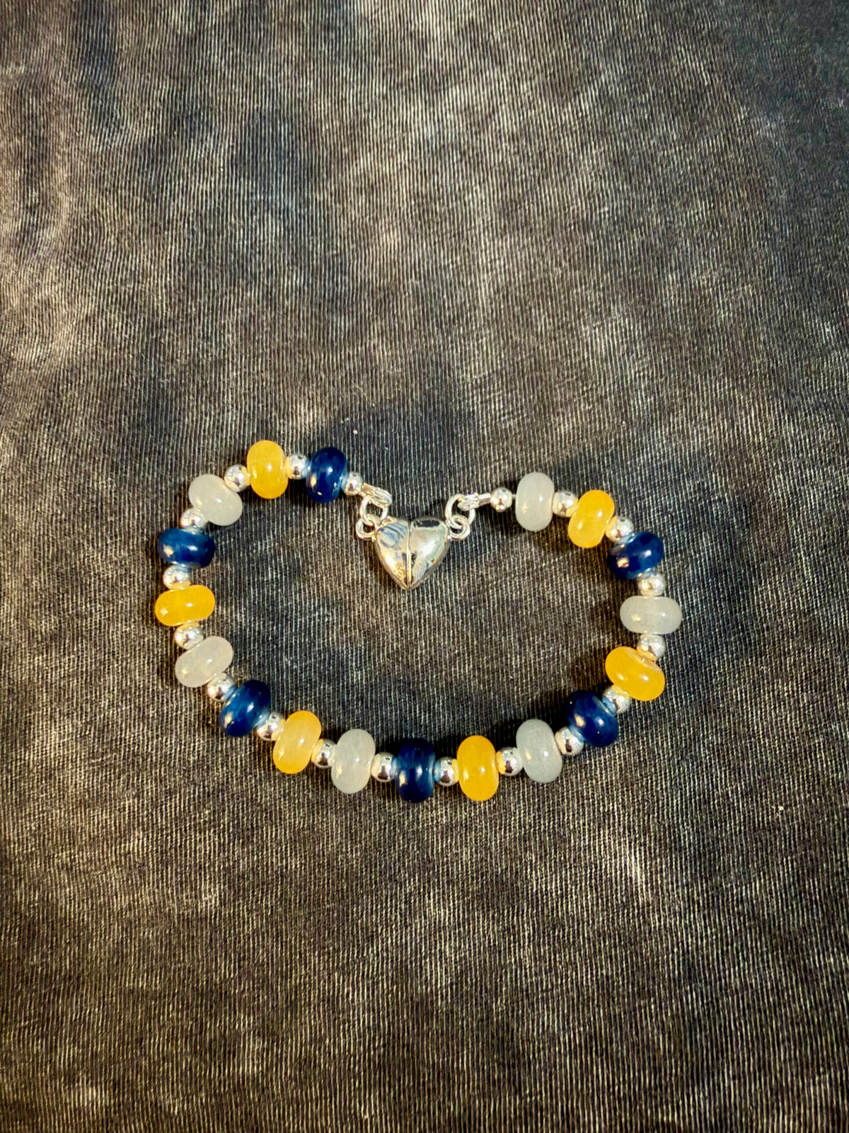 A beaded stretch bracelet featuring navy, gold, and white beads with a silver heart clasp, displayed on a textured gray background.