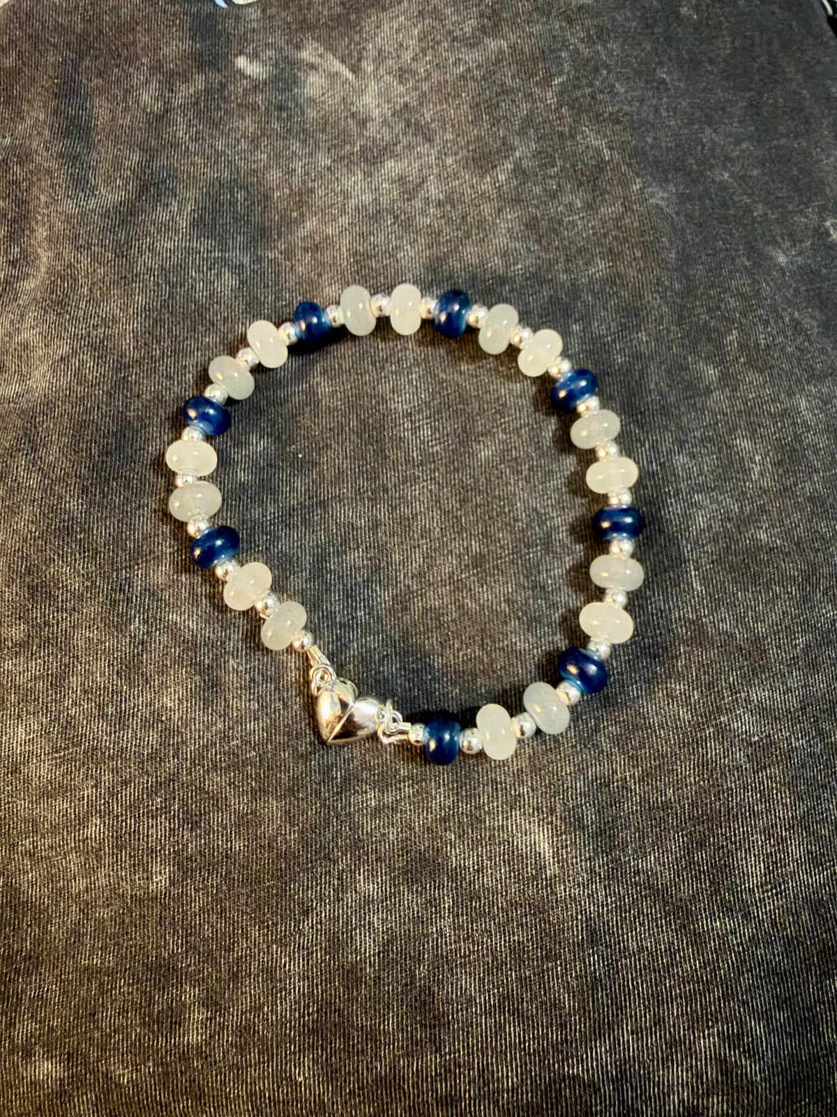 A beaded bracelet featuring milky white beads, deep blue beads, smokey grey beads, and a small silver-toned heart shaped clasp, displayed on a dark textured fabric background.