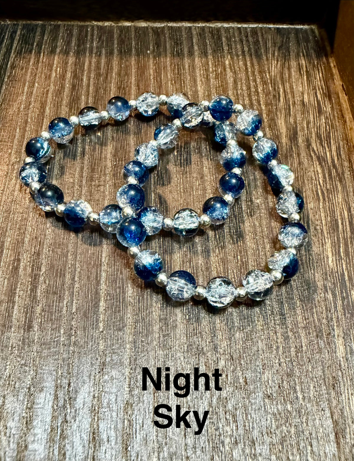 Two crackle glass beaded stretch bracelets with blue and clear crackle glass beads and silver spacer beaded, displayed on a dark wood surface with the text 'Night Sky' below.