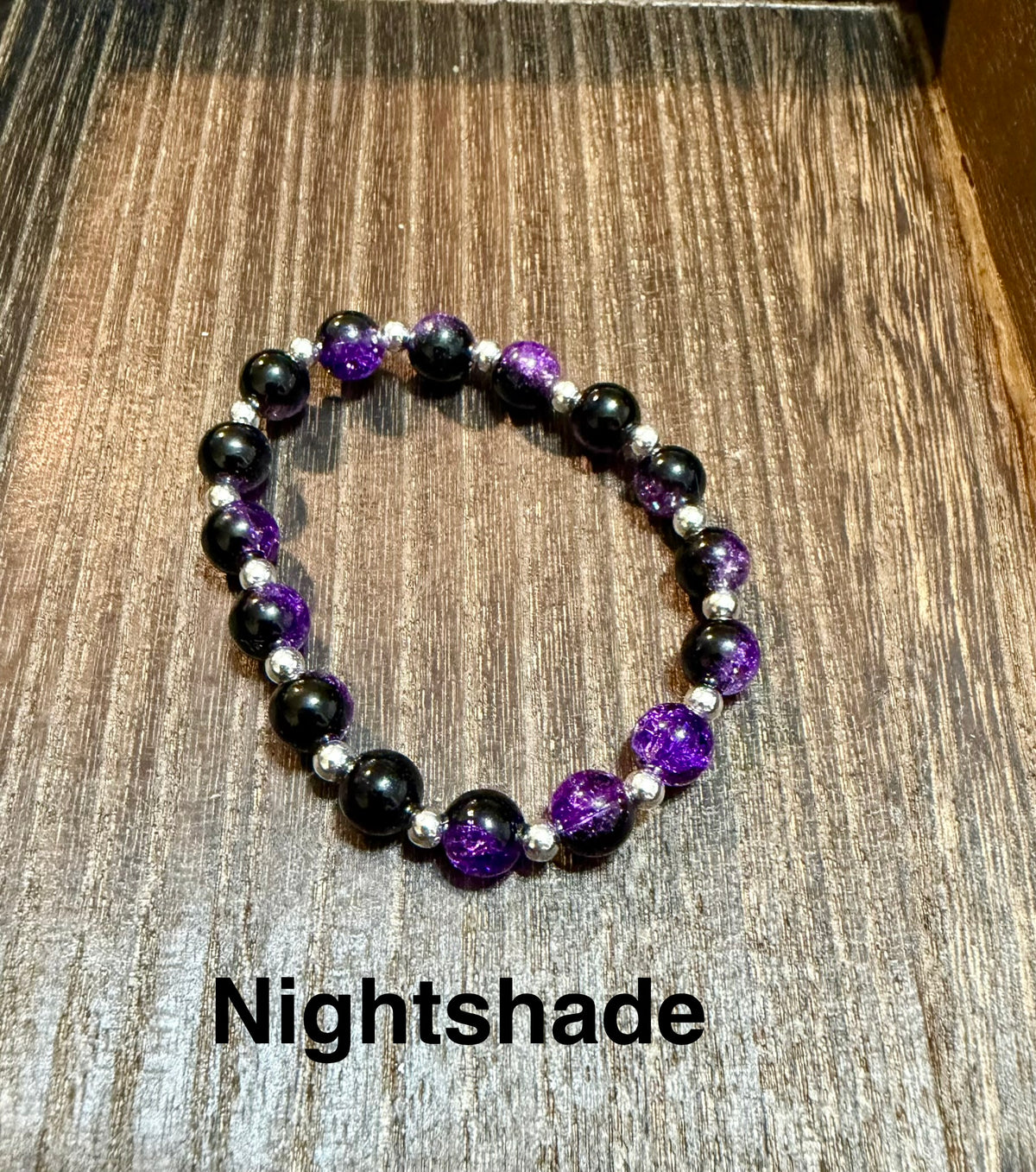 A beaded stretch bracelet with black and purple glittery crackle glass beads separated by small silver spacer beads, displayed on a wooden surface with the text 'Nightshade' underneath.