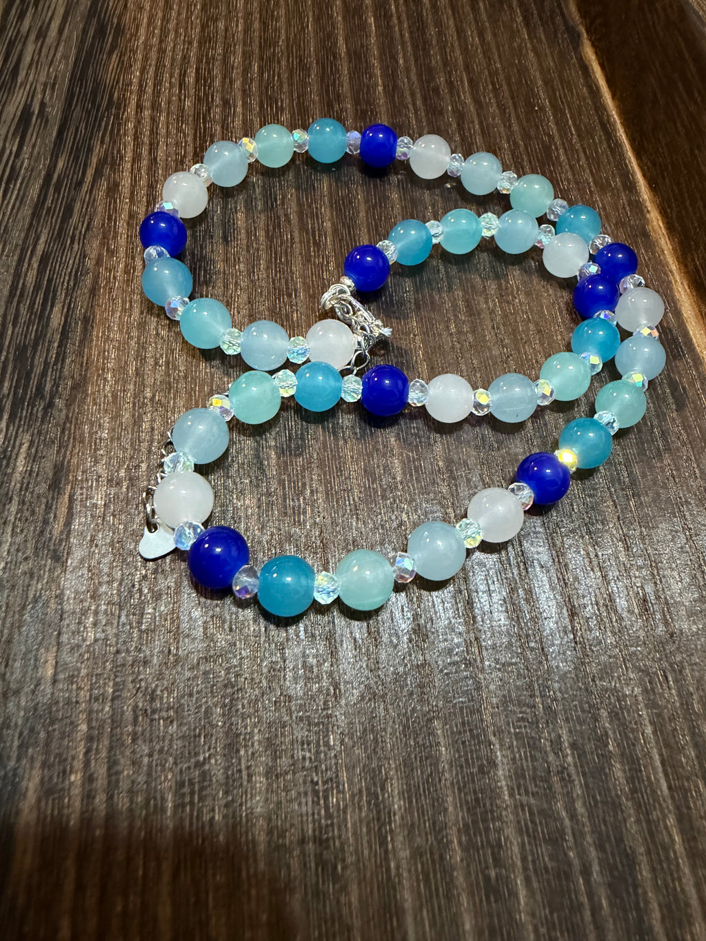 A beaded necklace with white, sky blue, light teal, light turquoise, and royal blue round glass beads, aurora borealis Austrian glass faceted spacer beads, and a heart dangle extender chain, displayed on a dark wood surface.